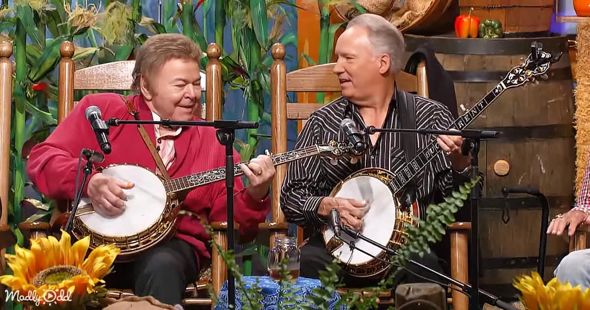Two men play banjos on a stage with microphones, surrounded by corn and flowers.