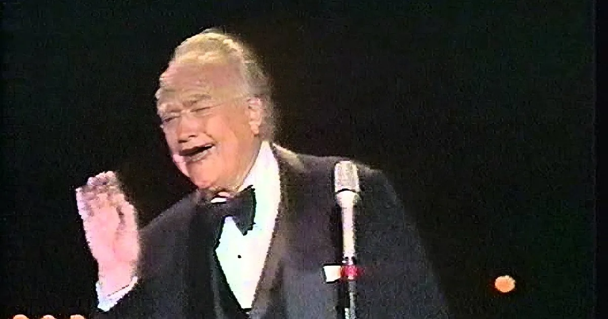A man in a tuxedo and bowler hat sings into a microphone on stage.