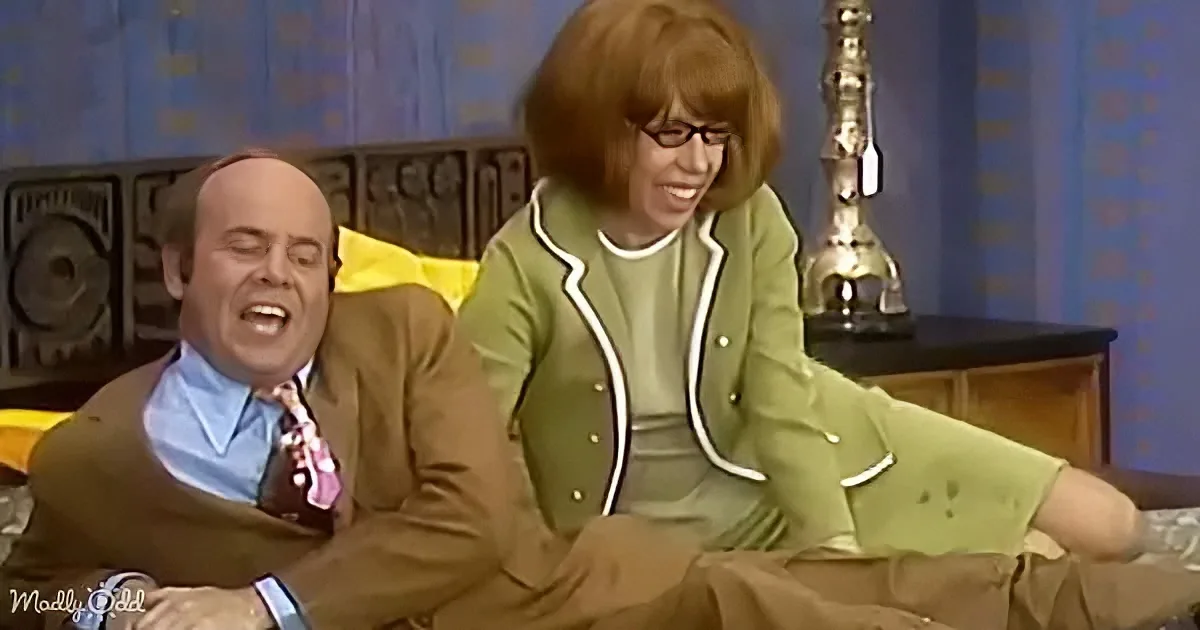 A man in a brown suit and a woman in a green outfit are sitting on a bed, laughing together.