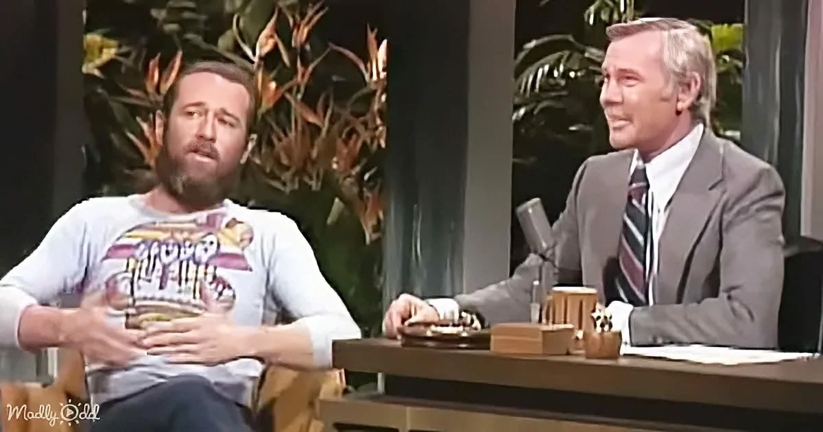 A man in a casual shirt sits with his hands on his stomach, facing another man in a suit at a desk.