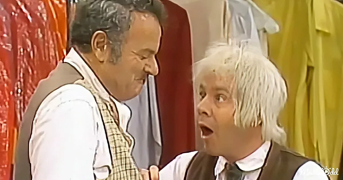 A man in a white shirt and vest stands next to another man with white hair, both in a room with colorful clothing.