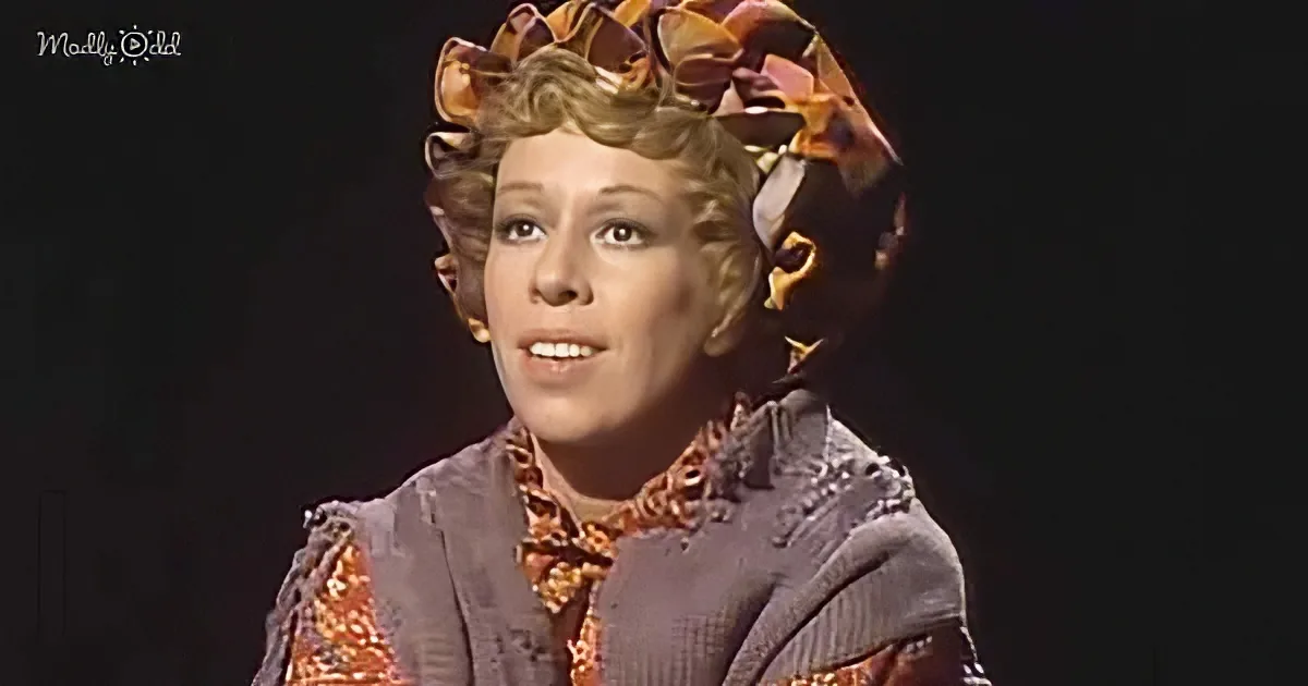 A woman in a vintage outfit with a floral hat and patterned blouse smiles against a dark background.