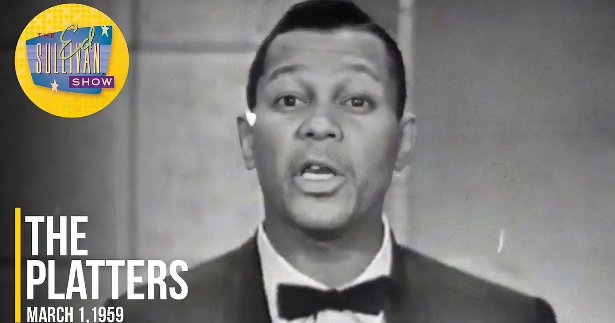 A man in a tuxedo sings passionately, surrounded by a group of musicians in a vintage setting.