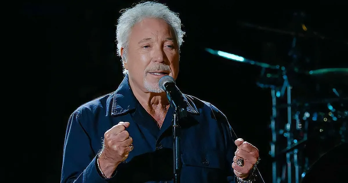 A man with white hair and a beard sings into a microphone, wearing a dark shirt, with his hands clenched.