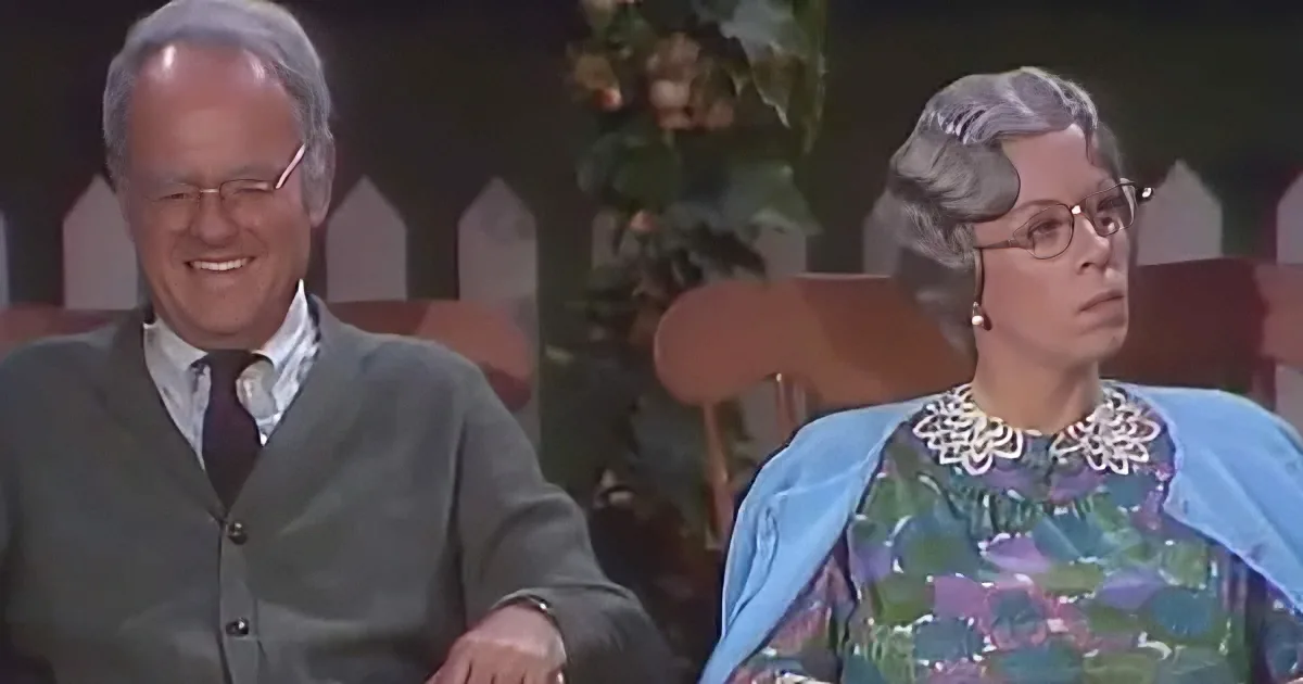 A man in a green sweater and tie smiles while a woman in a floral dress and blue shawl sits beside him.