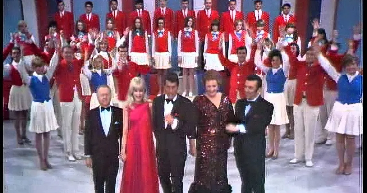 A group of performers in red and white costumes with a front row in formal attire, celebrating on stage.