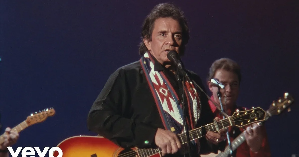 A man in a suit plays guitar on stage, singing into a microphone, accompanied by other musicians.