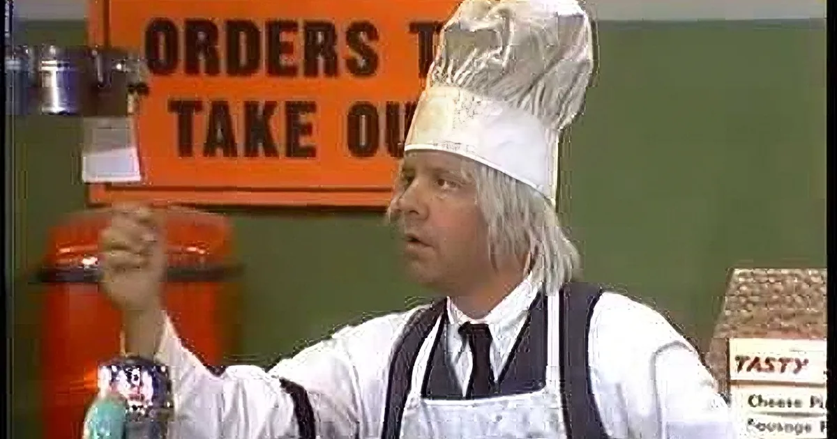 A man in a white shirt and suspenders gestures animatedly while sitting at a diner counter.
