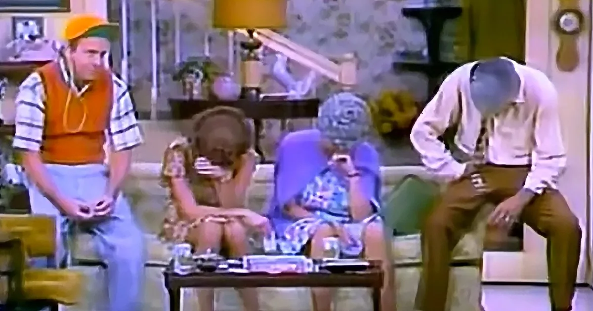 A man in an orange vest stands while others sit on a couch, appearing distressed or contemplative.