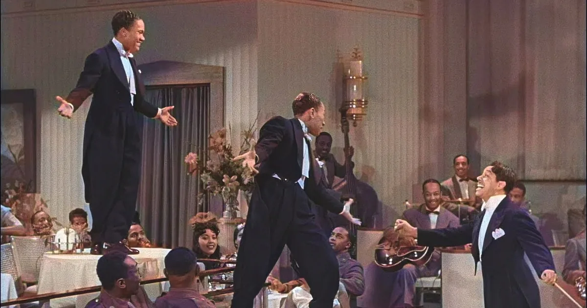 A man in a suit dances energetically in a room filled with people, some seated at tables.