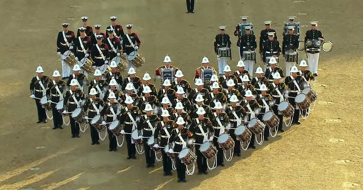 A marching band in uniform stands in formation, holding drums, with a few individuals in the background.