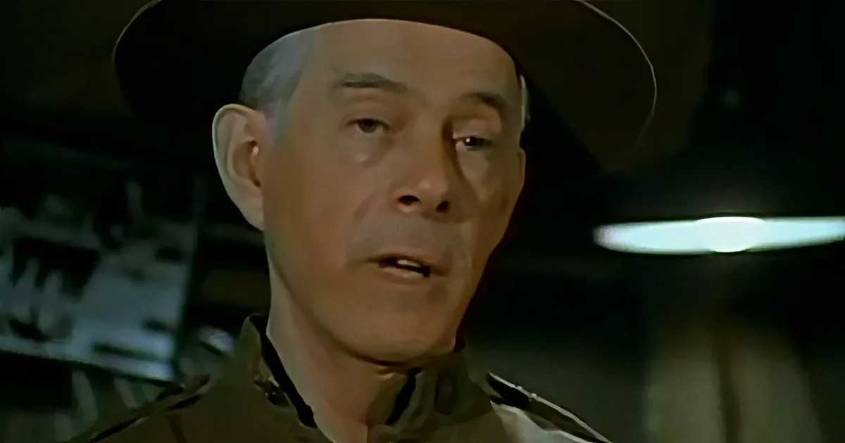 A man in a military uniform and hat appears to be speaking, with a dimly lit background.
