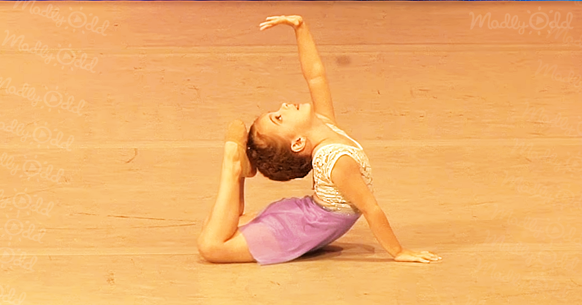 A young dancer in a purple skirt and white top performs a stretch on a wooden floor.