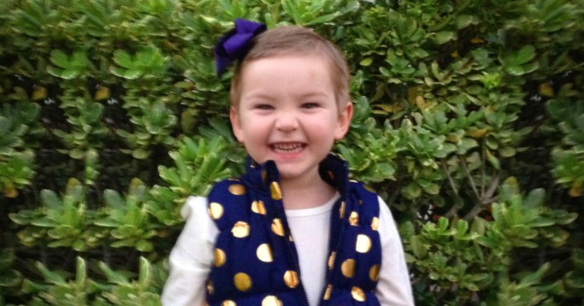 A young child with a purple bow, wearing a blue polka dot vest over a white shirt, smiles in front of a green bush.