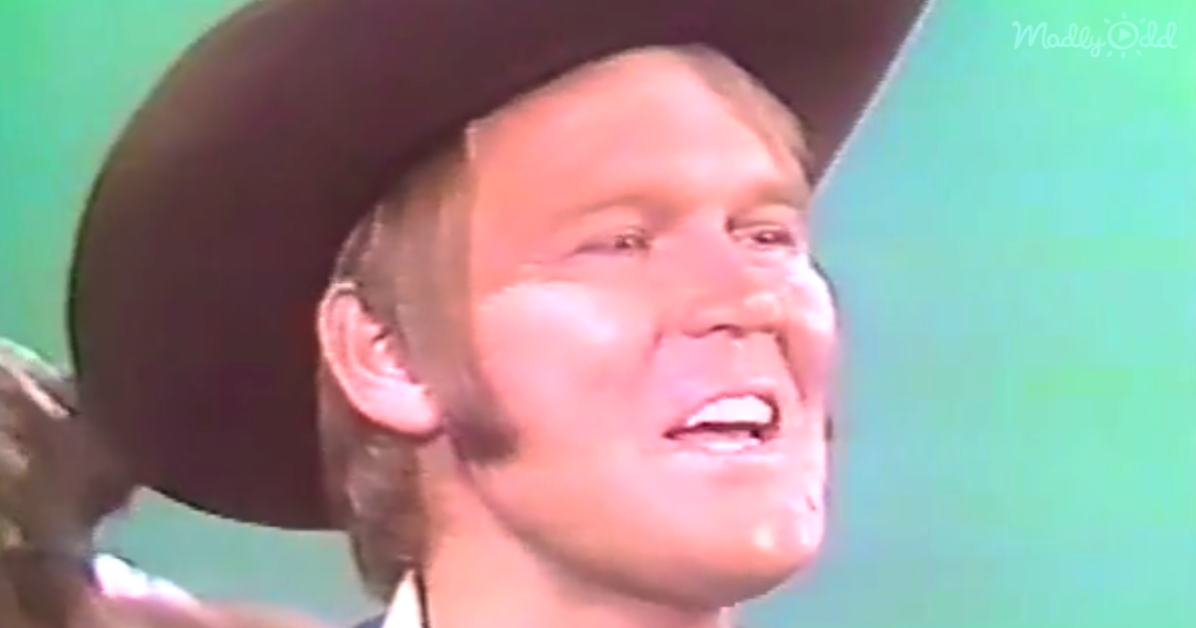 A man wearing a cowboy hat is singing with his eyes closed, mouth open, and a green background.