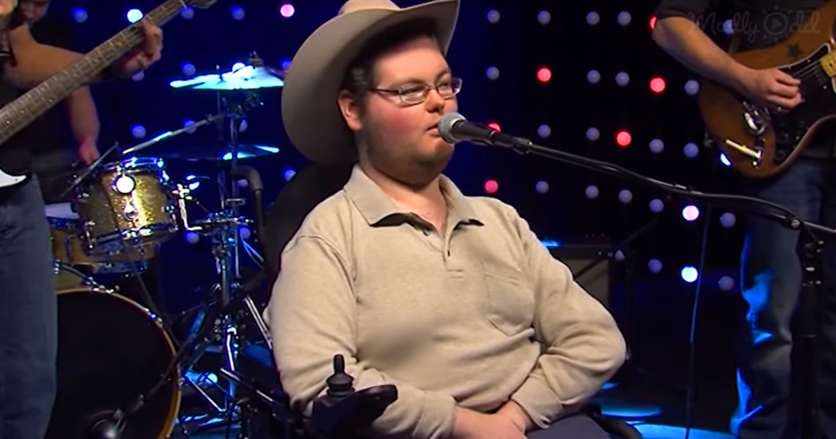 A man in a cowboy hat sings into a microphone, surrounded by a band and colorful lights.