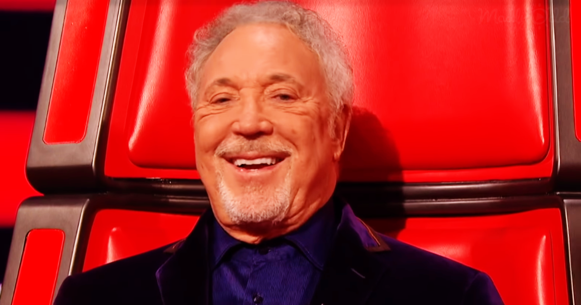A smiling man with gray hair and a beard, wearing a dark blue shirt, sits in a red chair.