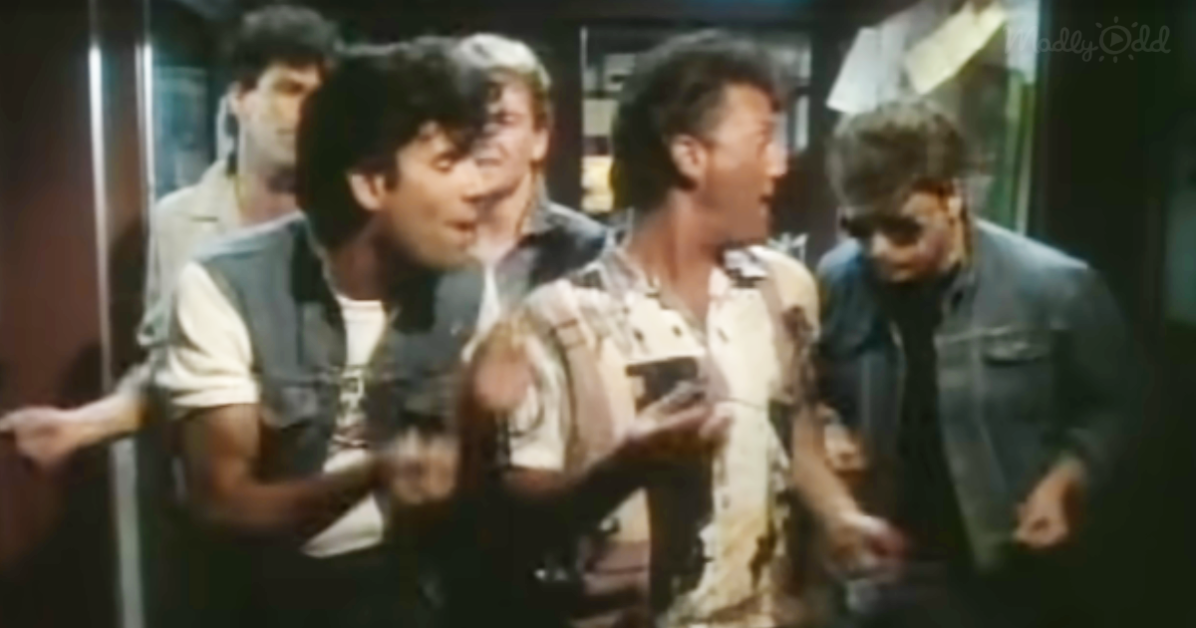 A man in a white shirt and vest is holding a phone, surrounded by others in a crowded, dimly lit setting.