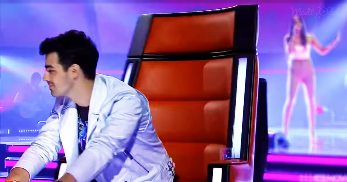 A man in a white jacket leans forward, watching a singer on stage with a red chair in the foreground.